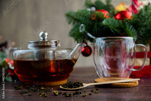 A transparent teapot with tea stands on the table, a transparent mug stands on a wooden stand next to it, a wooden spoon lies next to it and tea is scattered on a dark background. High quality photo
