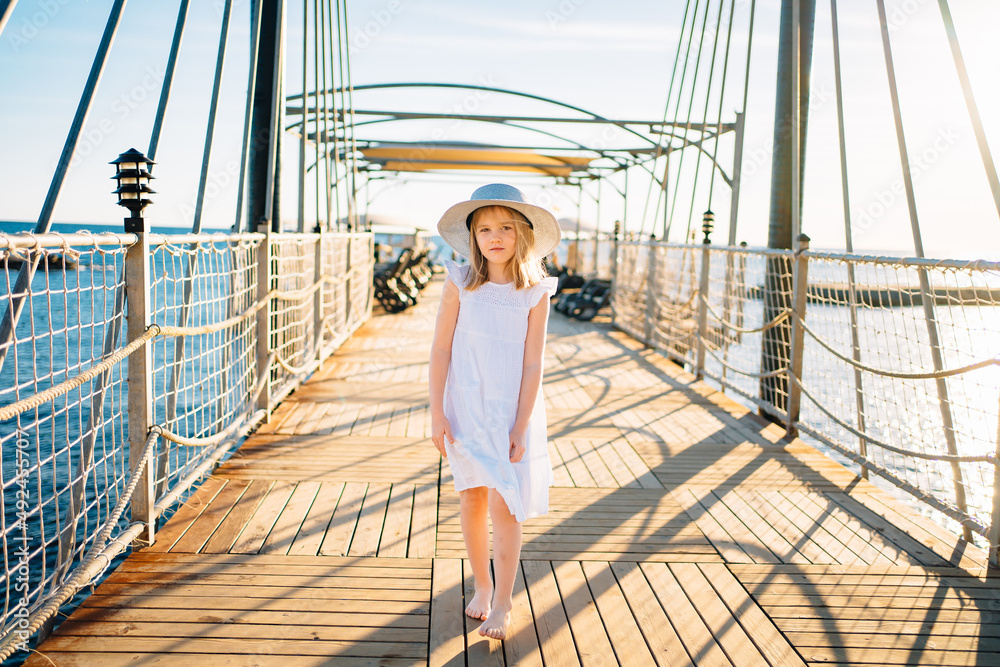 a little girl blonde in a white dress and a wide-brimmed hat stands on the pier