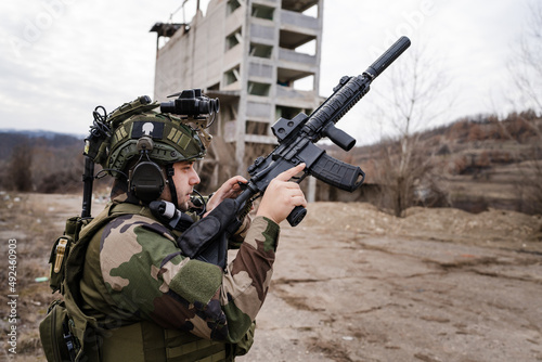One man soldier armed with rifle wearing camouflage uniform loading gun while standing in front of ruined building in battle zone dogs of war professional mercenary or volunteer side view copy space