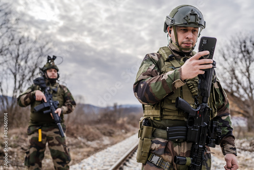 Two soldiers dogs of war mercenaries men in uniform armed service rifles standing while securing the railroad in combat zone in war soldier use mobile phone while patrolling area during mission photo