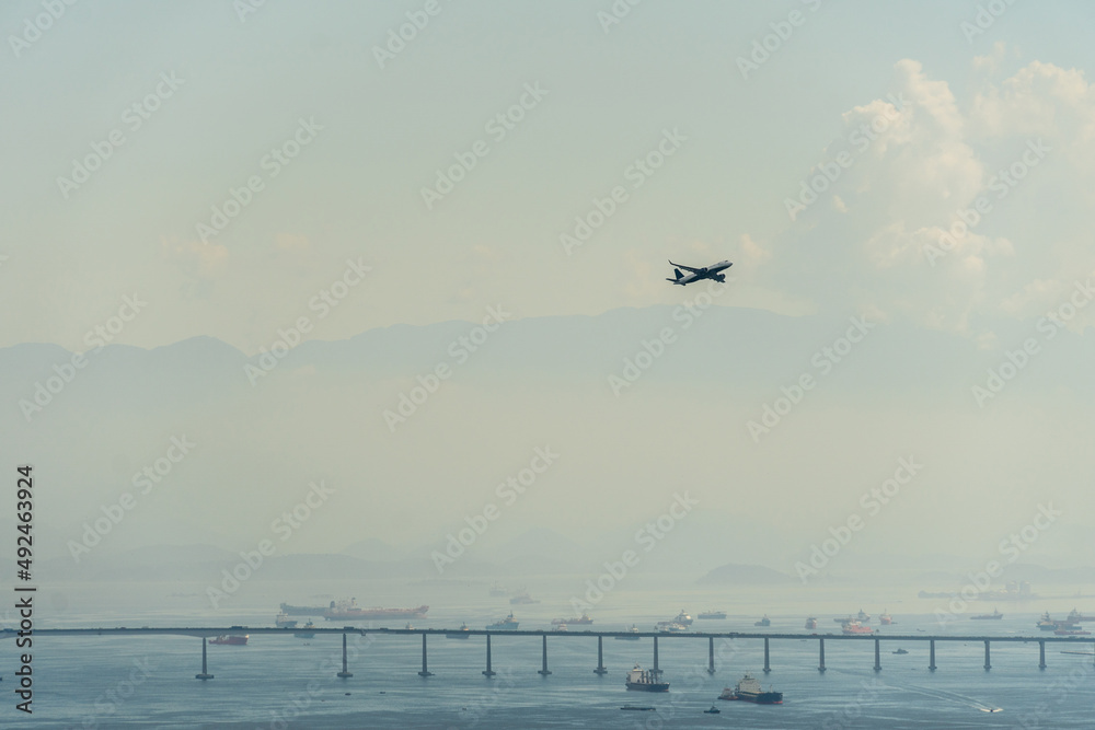 Airplane taking off from the airport runway in Rio de Janeiro, Brazil. Rio Niterói Bridge, mountains and ships in the background. Dawn with a little fog