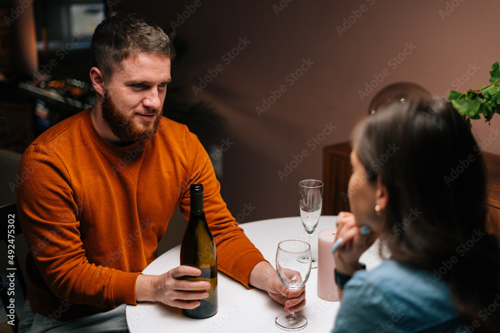 High-angle view of young man pouring wine into glass during talking with wife sitting together at festive table. Love couple celebrating anniversary or Valentine day having romantic dinner High-angle view of young man pouring wine into glass during talking with wife sitting together at festive table. Love couple celebrating anniversary or Valentine day having romantic dinner