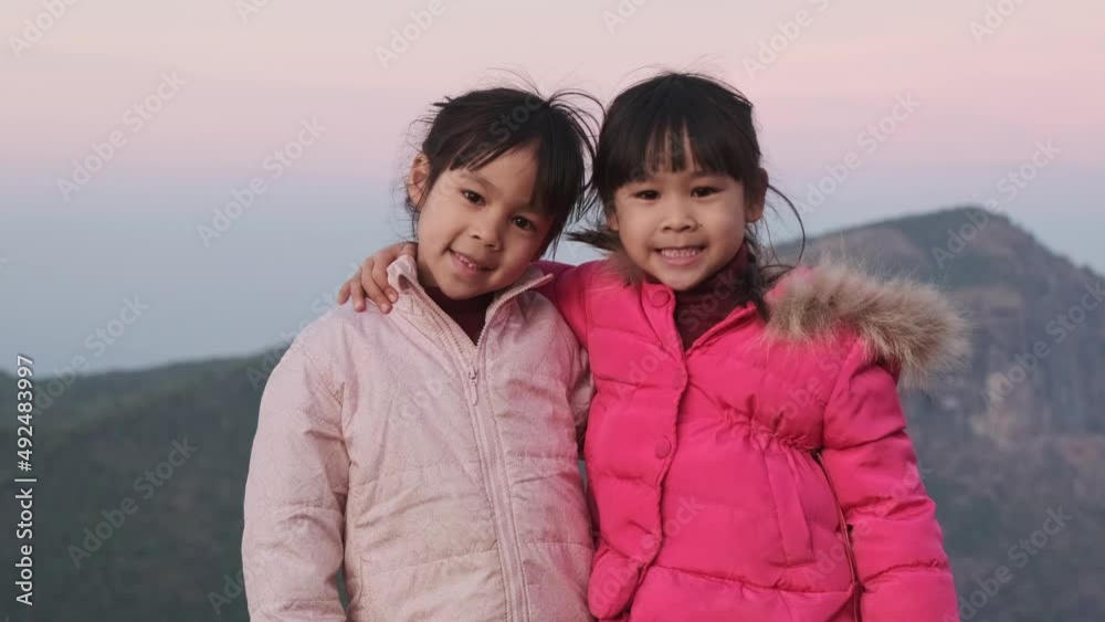 Two siblings little girls hugging on top of a mountain and looking at the camera against a sunset background. Hiking adventures with family on vacation. Two siblings little girls hugging on top of a mountain and looking at the camera against a sunset background. Hiking adventures with family on vacation.