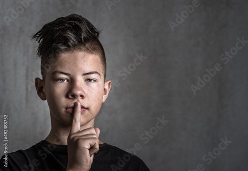 A teenage boy on a dark background pressed his index finger to his lips. Problems of adolescence in boys. No need to hush up the problems of puberty