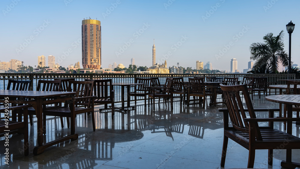 Morning in Cairo. On the terrace, on the banks of the Nile, there are tables and chairs. Reflection on the tiled floor. Buildings are visible on the far bank of the river. Blue sky. Egypt