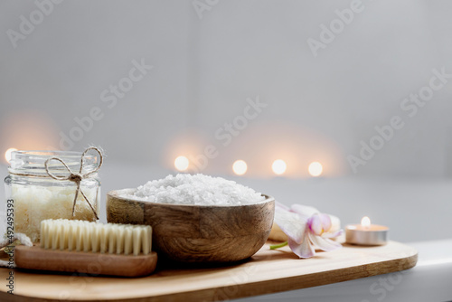 Preparation for hotel spa treatment or home bath procedure. White washbasin in bathroom with accessories on tray. Burning candles, washcloth, soap, foot brush, bottle with sea salt. Blurred background