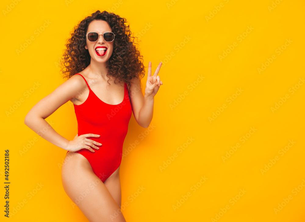 Young beautiful smiling woman posing near yellow wall in studio.Sexy model in red swimwear bathing suit.Positive female with curls hairstyle.Happy and cheerful. In sunglasses. Showing peace sign Young beautiful smiling woman posing near yellow wall in studio.Sexy model in red swimwear bathing suit.Positive female with curls hairstyle.Happy and cheerful. In sunglasses. Showing peace sign
