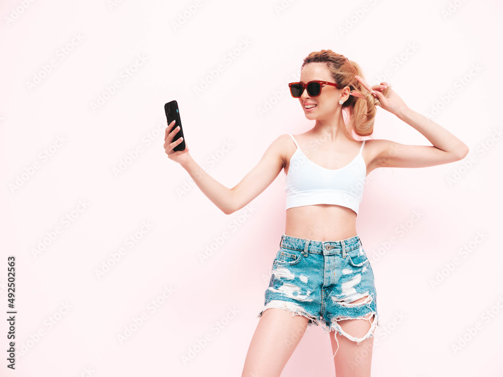 Young beautiful smiling female in trendy summer jeans shorts. Sexy carefree blond woman posing near wall in studio. Positive model having fun. Cheerful and happy in sunglasses. Isolated.Taking selfie Young beautiful smiling female in trendy summer jeans shorts. Sexy carefree blond woman posing near wall in studio. Positive model having fun. Cheerful and happy in sunglasses. Isolated.Taking selfie