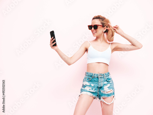 Young beautiful smiling female in trendy summer jeans shorts. Sexy carefree blond woman posing near wall in studio. Positive model having fun. Cheerful and happy in sunglasses. Isolated.Taking selfie