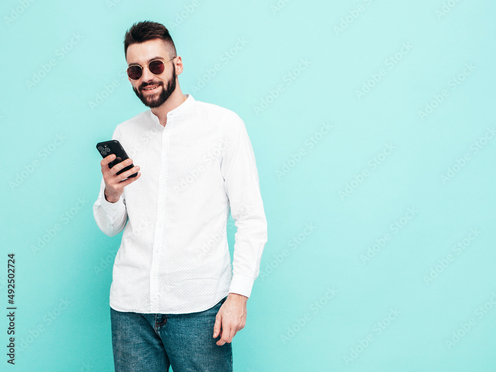 Handsome confident model.Sexy stylish man dressed in shirt and jeans. Fashion hipster male posing near blue wall in studio. Holding smartphone. Looking at cellphone screen. Using apps. Isolated Handsome confident model.Sexy stylish man dressed in shirt and jeans. Fashion hipster male posing near blue wall in studio. Holding smartphone. Looking at cellphone screen. Using apps. Isolated