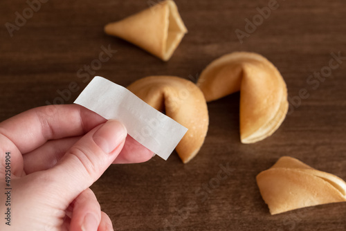 Person holds in hand blank paper slip from fortune cookie against few cookies laying on table surface background, mockup for your good luck wish