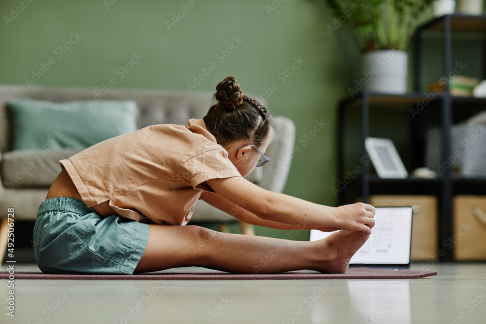 Obraz premium Side view portrait of teenage girl doing stretching exercises while enjoying yoga at home via online lesson