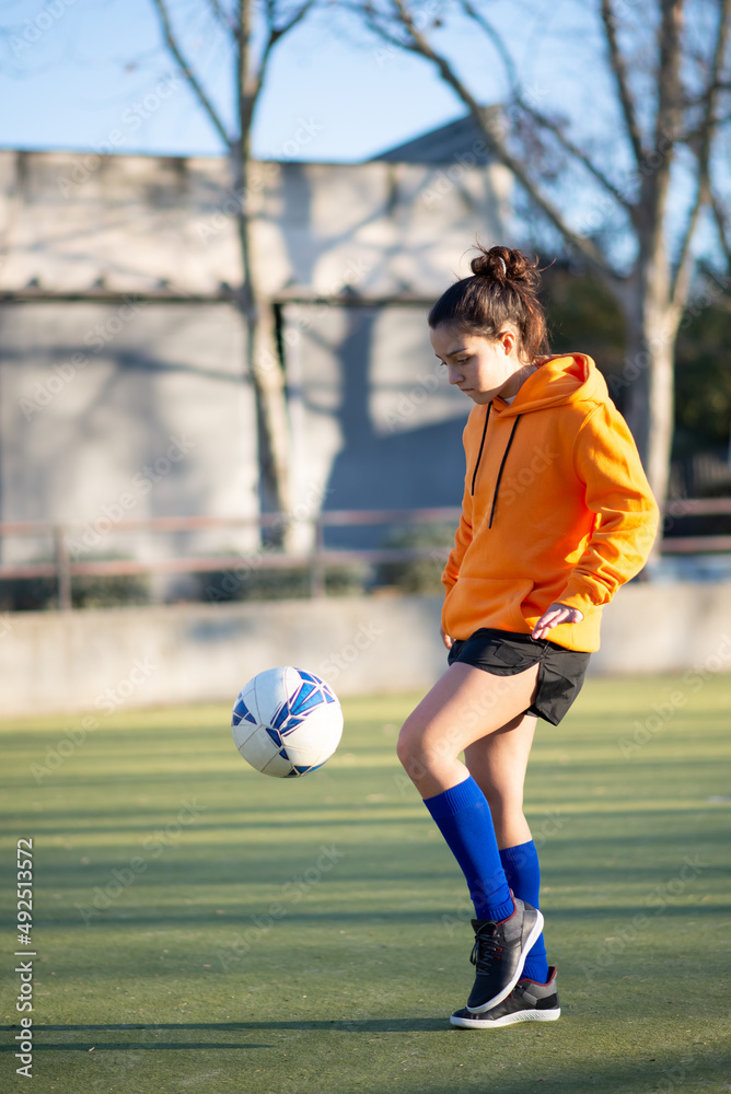 Young girl training playing ball on field. Beautiful Caucasian girl in sportswear standing on green field concentrated on kicking ball. Healthy lifestyle and sport concept