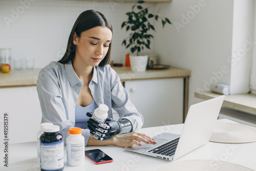 Healthy lifestyle. Pretty female in shirt sitting at kitchen table in front of laptop and food supplements in bottles, holding one jar of vitamins in bionic prosthetic black metal hand, making order photo