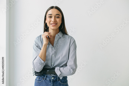 Horizontal portrait of female in jeans and striped shirt with round earrings and smooth long hair posing against white wall with copy space for your advertising content, having bionic prosthetic hand