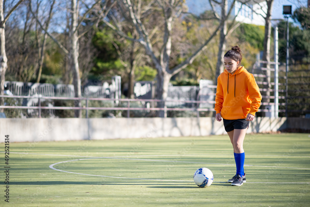 Portrait of teenage girl with ball on green field. Concentrated girl in sportswear playing football with her team standing in center of field ready for kicking. Healthy lifestyle, team sport concept
