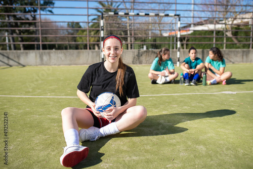 Portrait of smiling teenage football girl on field. Happy girl in sportswear resting on ground looking at camera while other girls sitting behind her enjoying talk. Active rest and team sport concept