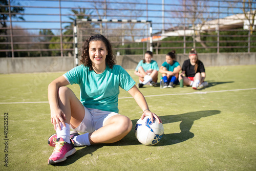 Portrait of happy female football player posing on field. Beautiful girl sitting with ball looking at camera and other girls resting behind, communicating. Hobby, leisure activity, team sport concept © KAMPUS
