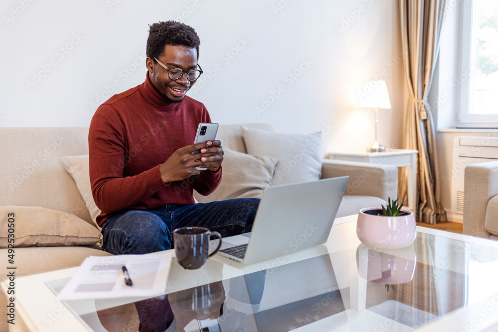 Excited Black African American Man Having a Video Call on Smartphone while Sitting on a Sofa in Living Room. Happy Man Smiling at Home and Talking to His Friends and Family Over the Internet.....