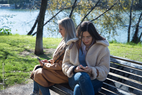 Real couple of young women, sitting on a bench, back to back and checking social networks on their cell phones. Concept lgtbiq+, lesbians, smartphone, visibility.