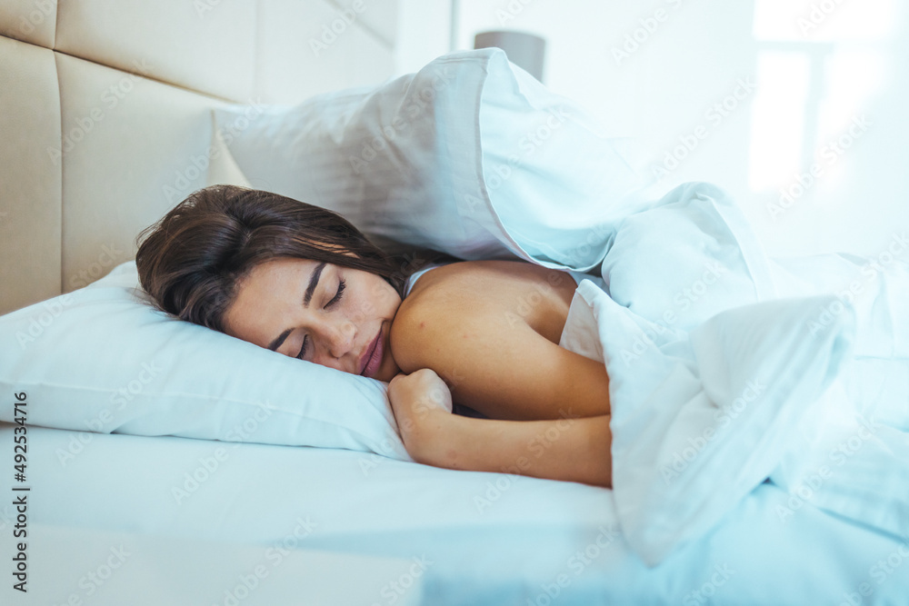 Horizontal shot of relaxed carefree female under white bedclothes in bed at bedroom, sees pleasant dreams, keeps eyes closed, enjoys good rest at home and calm atmosphere. People and rest concept Horizontal shot of relaxed carefree female under white bedclothes in bed at bedroom, sees pleasant dreams, keeps eyes closed, enjoys good rest at home and calm atmosphere. People and rest concept