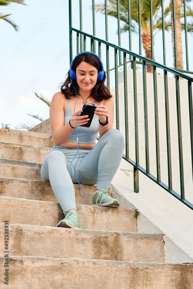 Woman in sportswear sitting looks at her smartphone and listens to music
