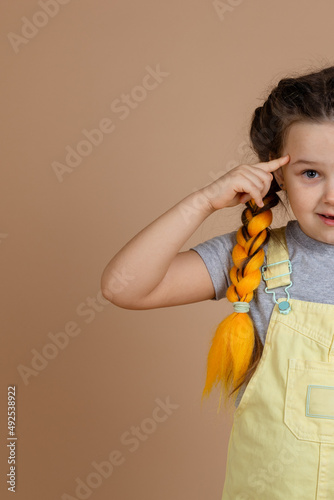 Half of surprised young girl, with yellow kanekalon pigtails looking at camera holding finger by temple in yellow jumpsuit and gray t-shirt on beige background.
