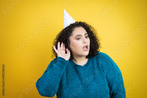 Young beautiful woman wearing a birthday hat over isolated yellow background surprised with hand over ear listening an hearing to rumor or gossip © Irene