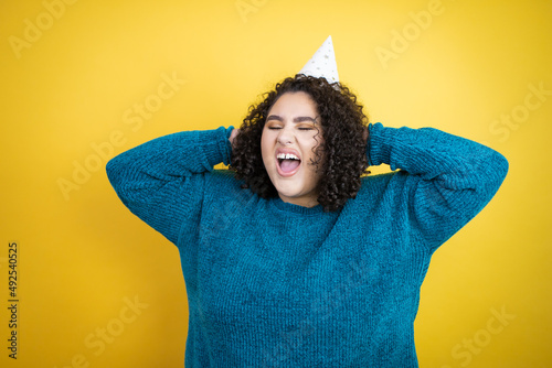 Young beautiful woman wearing a birthday hat over isolated yellow background relaxing and stretching, arms and hands behind head and neck smiling happy © Irene