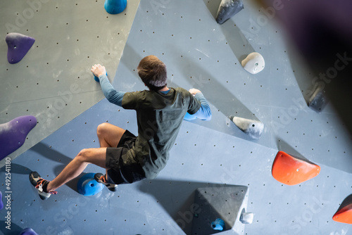 Back view of strong athlete training climbing artificial wall. Caucasian guy doing bouldering in gym holding wall with hands climbing up and balancing body. Active life and sport activity concept