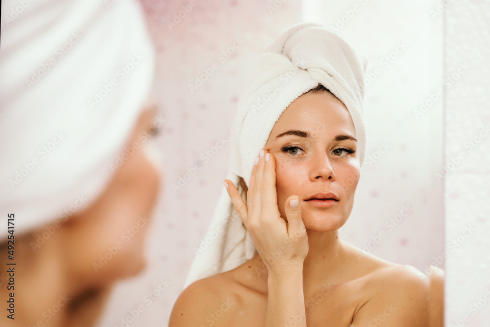 Young beautiful woman using face cream moisturizing lotion after bath. Pretty attractive girl in a towel on her head stands in front of a mirror in a home bathroom. Daily hygiene and skin care Young beautiful woman using face cream moisturizing lotion after bath. Pretty attractive girl in a towel on her head stands in front of a mirror in a home bathroom. Daily hygiene and skin care
