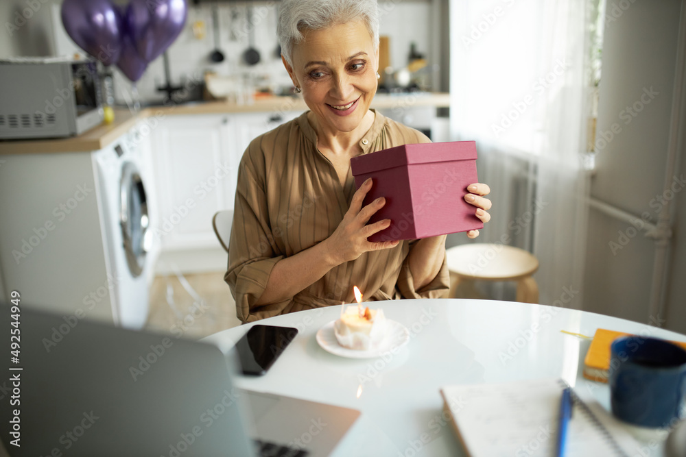 Attractive senior grey haired woman in light brown dress sitting at kitchen table with present box in hand, having online celebration via laptop web camera, smart phone and cupcake on table