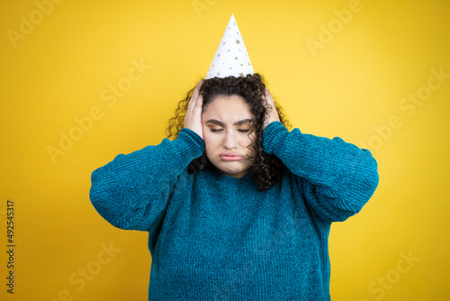 Young beautiful woman wearing a birthday hat over isolated yellow background thinking looking tired and bored with hands on head © Irene
