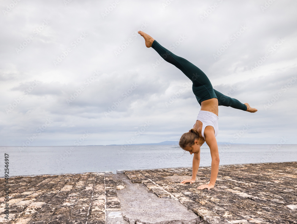Outdoor yoga practice near the ocean. Young woman practicing Adho Mukha  Vrksasana. Yoga Handstand is an inverted asana. Beautiful asana. Strong  slim body. Yoga retreat. Copy space. Bali Stock Photo