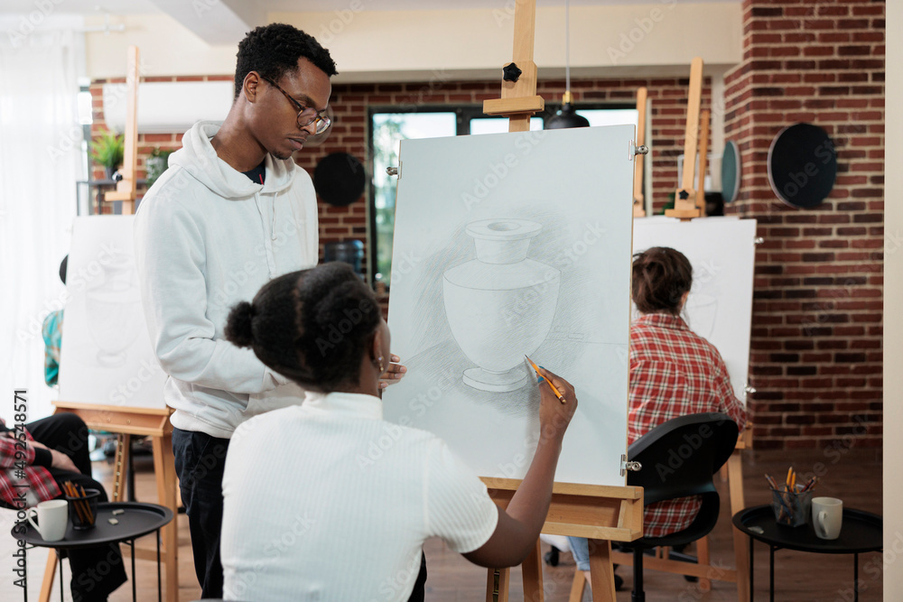 Painter teacher explaining illustration technique to student working together at graphic technique for school projct. Diverse group of artists drawing vase model on white canvas during art lesson