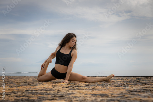 Sportive and athletic young female doing leg-split. Stretching exercise on the beach. Sport outdoors concept. Healthy lifestyle. Copy space. Caucasian woman wearing black sportswear. Bali © Olga