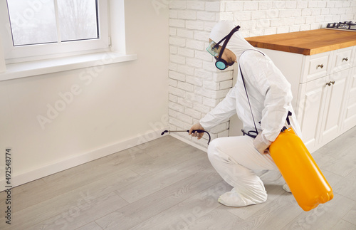 Pest control guy taking care of all the cockroaches or termites in the house. Male exterminator in white protective work uniform crouching by kitchen wall and spraying insecticide from sprayer bottle