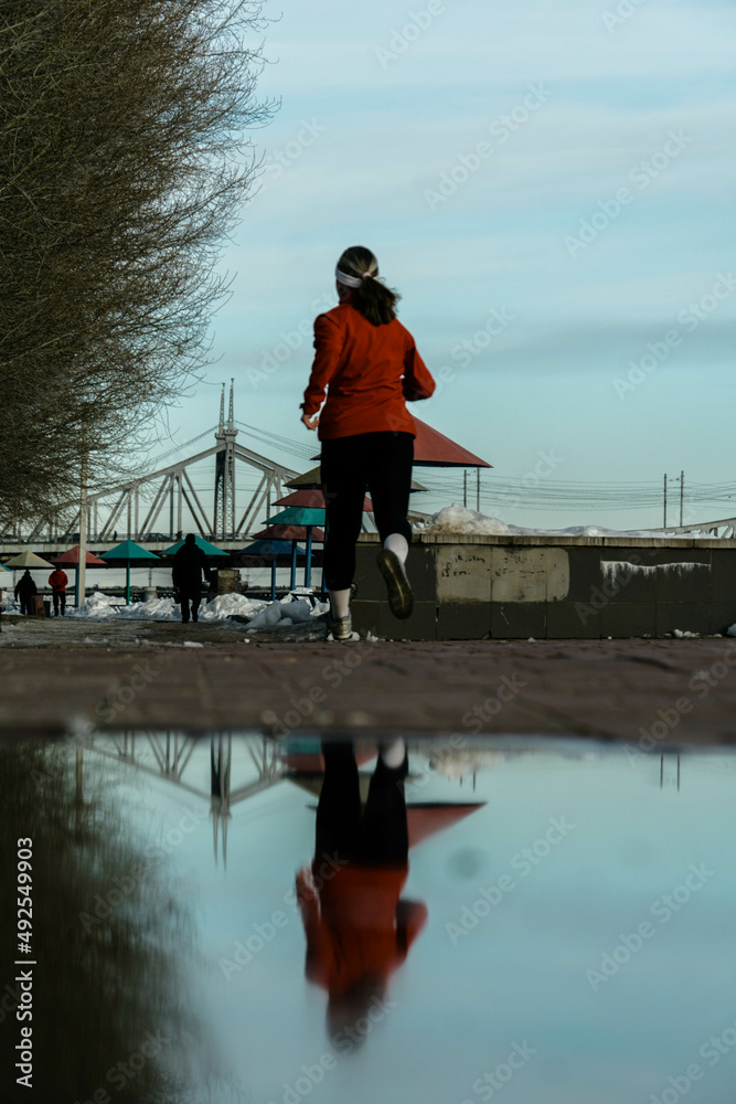 
A woman runs on the street, on the river bank, reflected in a puddle, photo in the afternoon