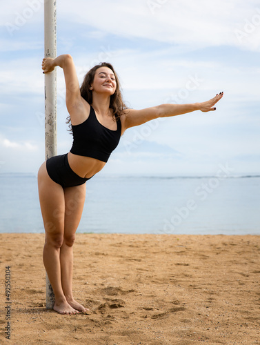 Attractive pole dance woman posing on the beach. Slim body. Young Caucasian woman wearing black sportswear. Fitness, wellness concept. Ocean and sky background. Copy space. Bali