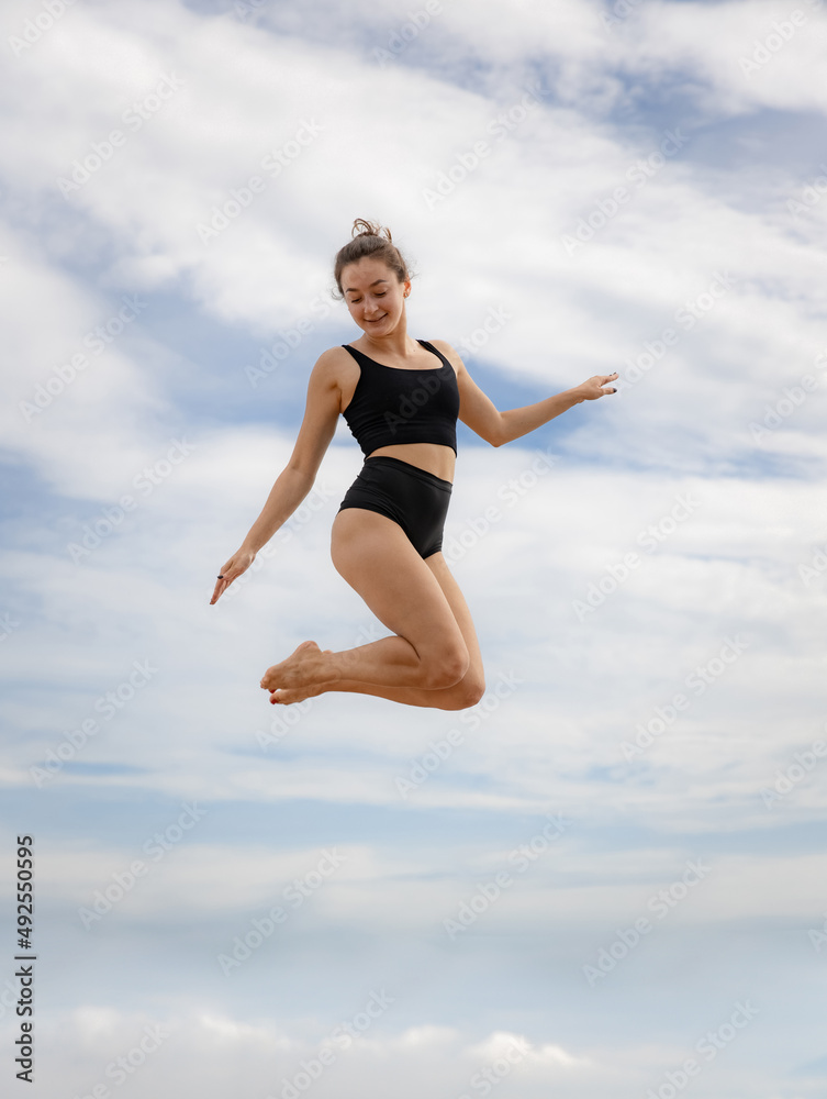 Beautiful gymnast woman jumping over cloudy blue sky. Caucasian woman wearing black sportswear. Fitness, wellness concept. Outdoor activity. Copy space. Sky background. Bali