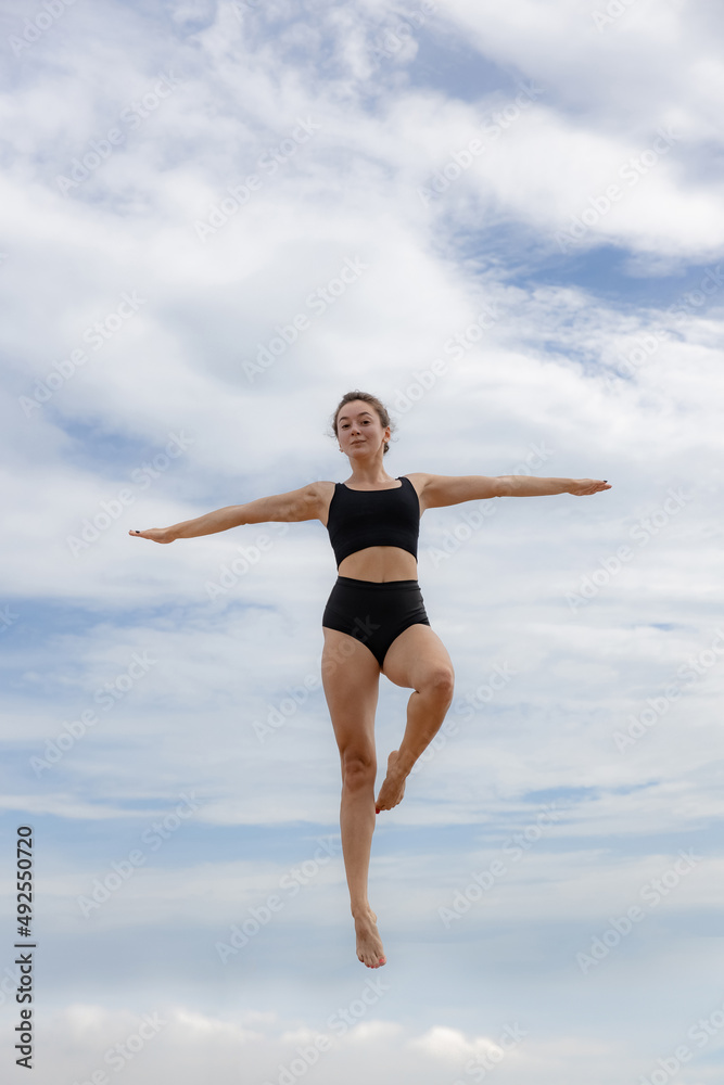 Attractive young woman jumping over cloudy blue sky. Caucasian woman wearing black sportswear. Fitness, wellness concept. Outdoor activity. Copy space. Sky background. Bali