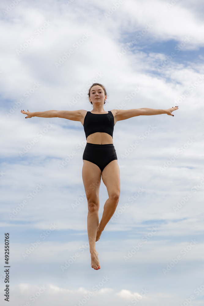 Beautiful young woman jumping over cloudy blue sky. Caucasian woman wearing black sportswear. Fitness, wellness concept. Outdoor activity. Copy space. Sky background. Bali Beautiful young woman jumping over cloudy blue sky. Caucasian woman wearing black sportswear. Fitness, wellness concept. Outdoor activity. Copy space. Sky background. Bali