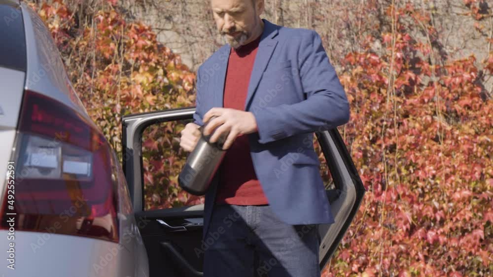 A middle-aged handsome Caucasian man pulls a vacuum bottle out of his car and drinks tea in fall