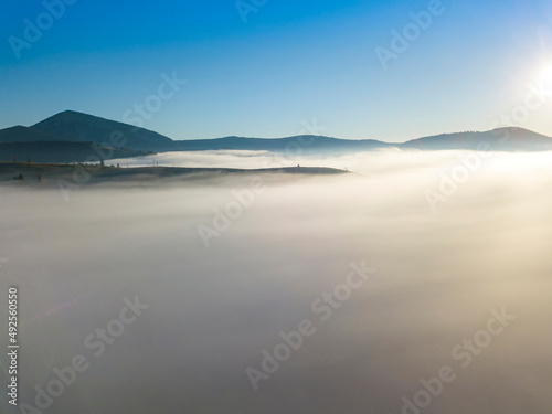 Flight over fog in Ukrainian Carpathians in summer. Mountains on the horizon. A thick layer of fog covers the mountains with a continuous carpet. Aerial drone view.