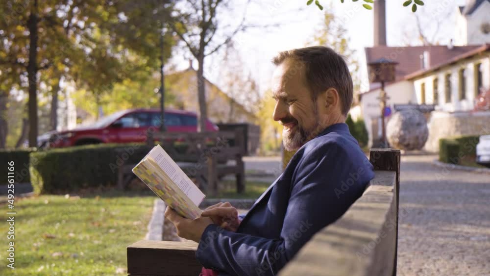 A middle-aged handsome Caucasian man reads a book with a smile as he sits on a bench in a quaint town in fall A middle-aged handsome Caucasian man reads a book with a smile as he sits on a bench in a quaint town in fall
