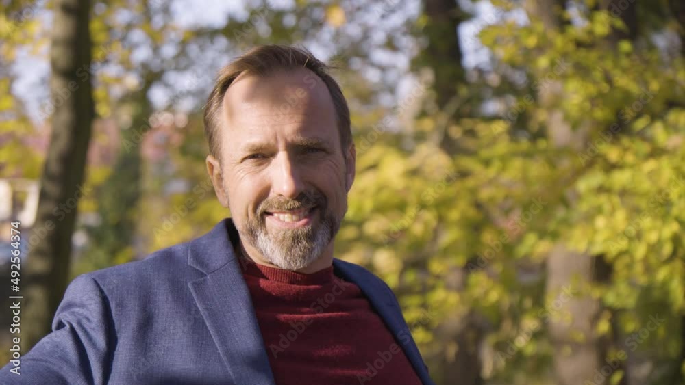 A middle-aged handsome Caucasian man smiles at the camera as he sits on a bench in a quaint town in fall A middle-aged handsome Caucasian man smiles at the camera as he sits on a bench in a quaint town in fall