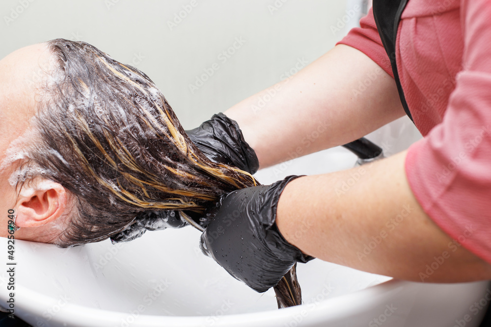 Naklejka premium The hairdresser in black gloves washing brunette woman's hair in the beauty salon.