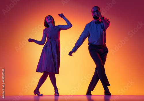 Portrait of excited man and woman, couple of dancers in vintage retro style outfits dancing lindy hop dance isolated on gradient yellow and purple background.