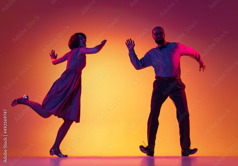 Portrait of excited man and woman, couple of dancers in vintage retro style outfits dancing lindy hop dance isolated on gradient yellow and purple background.