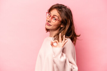Young caucasian woman isolated on pink background winks an eye and holds an okay gesture with hand.